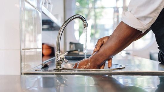 Pair of hands being washed under a running faucet in a restaurant or kitchen sink.